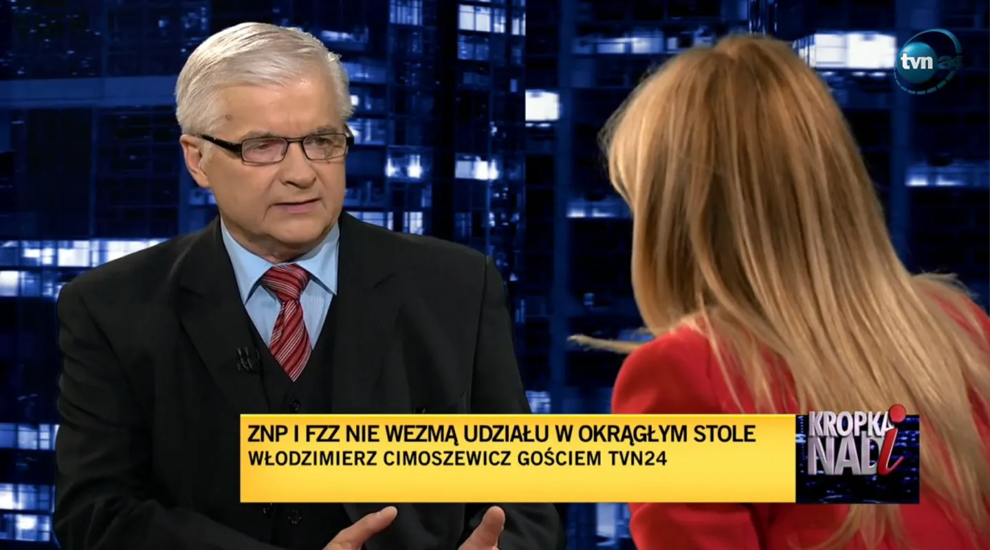 Wywiad w studiu telewizyjnym: starszy mężczyzna w garniturze i okularach rozmawia z kobietą o blond włosach, w tle niebieska panorama miasta, a na dole żółty pasek z napisem i logo TVN24.