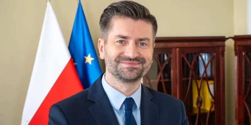 Uśmiechnięty mężczyzna w garniturze stoi obok flag Polski i Unii Europejskiej w gabinecie, w kontekście wyróżnienia MEP Award.