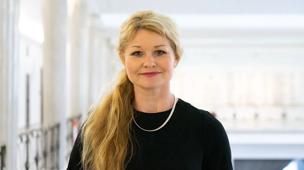 Uśmiechnięta kobieta o długich blond włosach związanych w koński ogon, ubrana w czarną bluzkę i srebrny naszyjnik, stoi w jasnym wnętrzu.