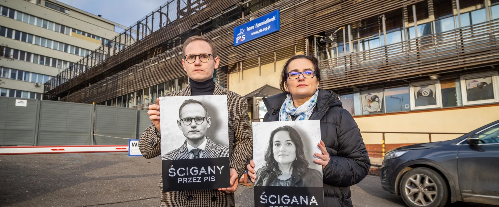 Dwóch dorosłych stoi na zewnątrz budynku, trzymając plakaty z czarno‑białymi portretami mężczyzny i kobiety oraz napisy „Ścigany przez PiS” i „Ścigana przez PiS”.