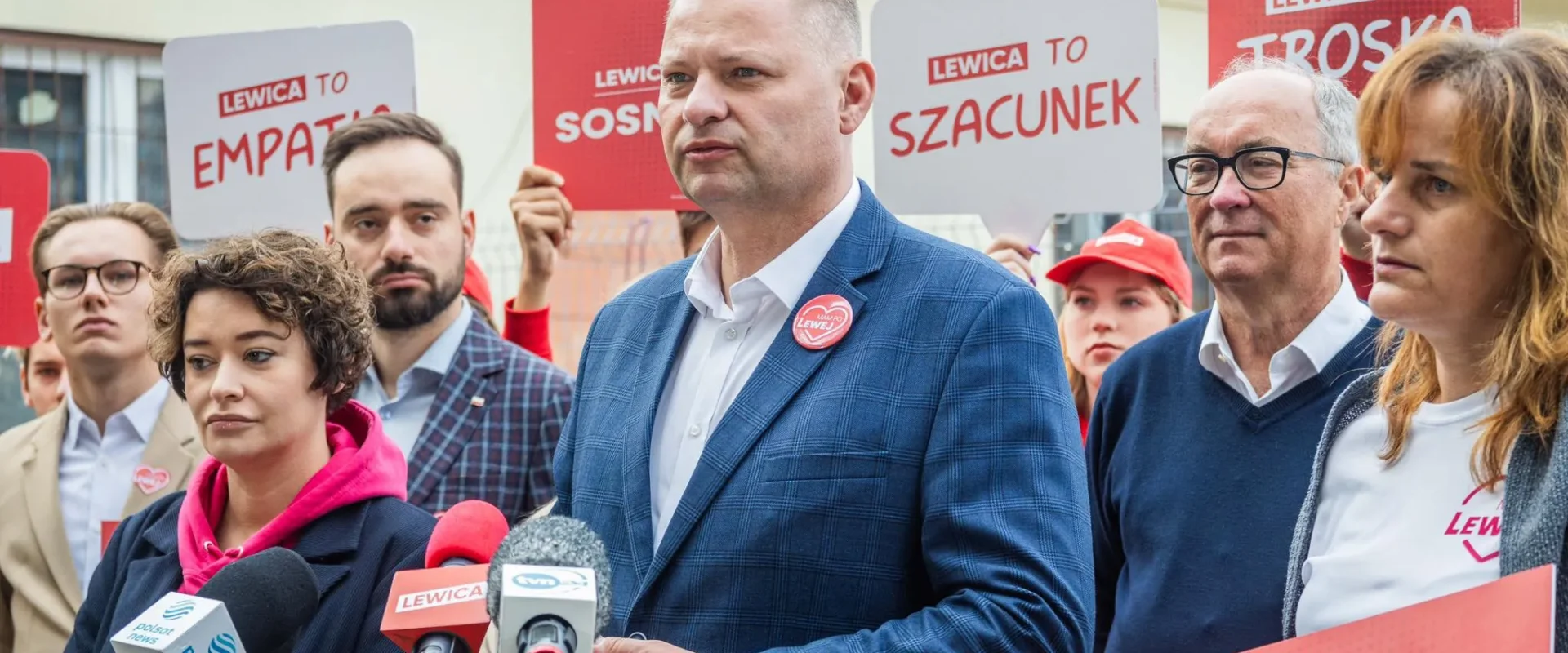 Grupa polityków Lewicy podczas konferencji prasowej, stojąca za mikrofonami, trzyma plakaty z hasłami „LEWICA TO EMPATIA”, „LEWICA TO SZACUNEK” i „LEWICA TO TROSKA”.