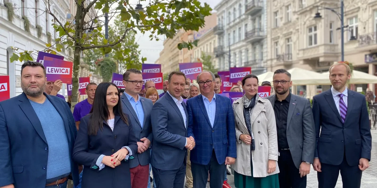 Grupa polityków Lewicy w marynarkach stoi na ulicy, w tle widoczne plakaty z napisem LEWICA, nawiązujące do zakończenia procesu konsolidacji przed kongresem zjednoczeniowym.