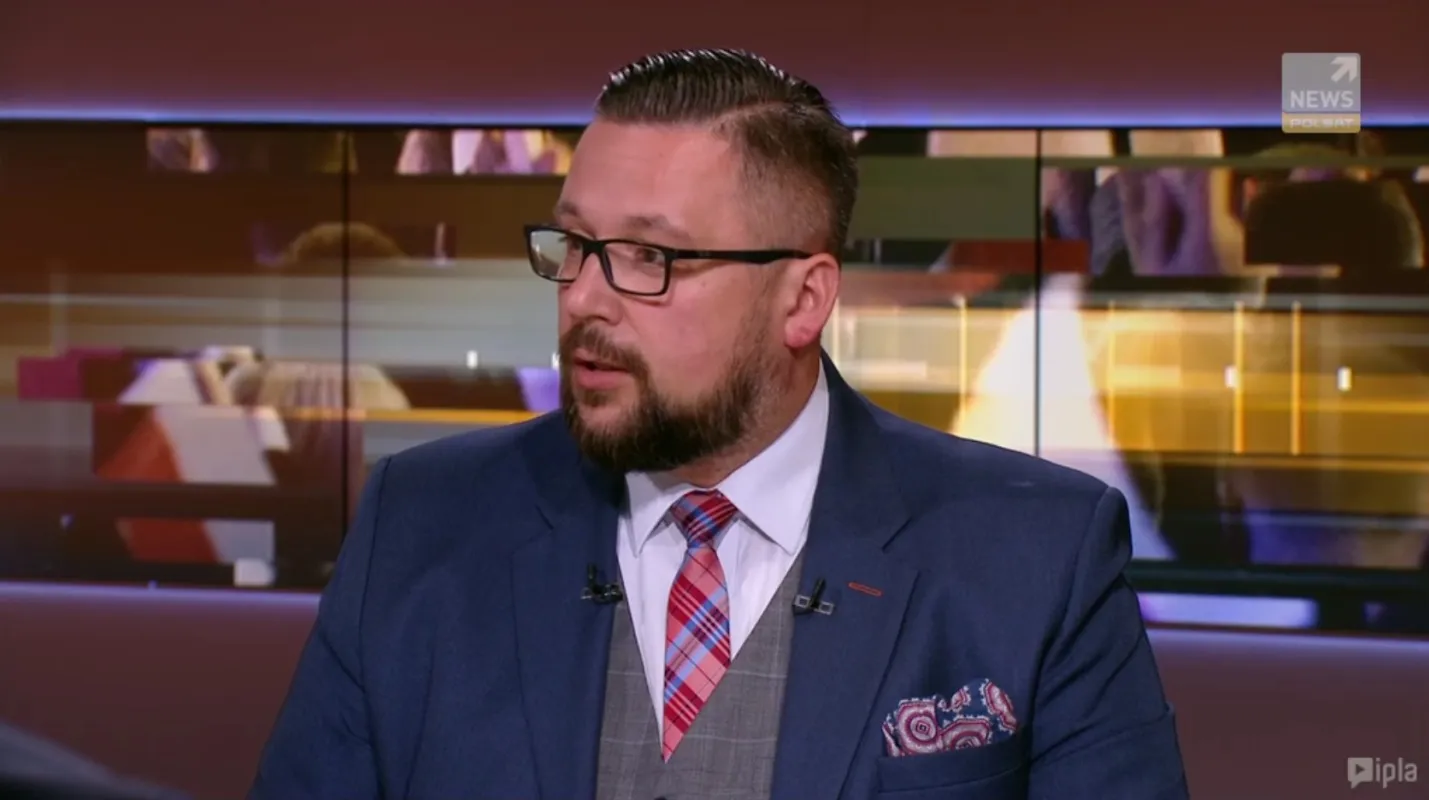Mężczyzna w granatowym garniturze, białej koszuli i różowo‑czerwonym kraciastym krawacie siedzi w studiu telewizyjnym z mikrofonami na klapach.