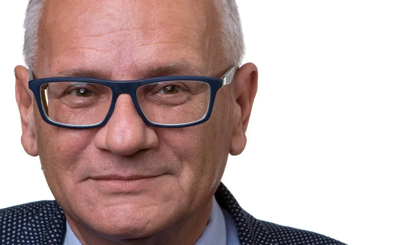 Portret starszego mężczyzny z siwymi włosami i okularami, ubrany w granatowy garnitur z drobnym wzorem i niebieską koszulą na białym tle.