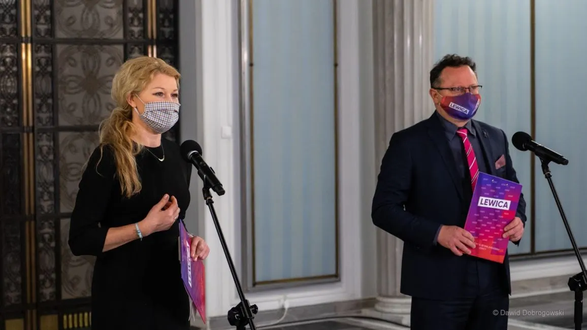 Dwóch polityków Lewicy w maseczkach stoi przy mikrofonach podczas konferencji prasowej, trzymając materiały z napisem Lewica, w tle kolumny i drzwi.