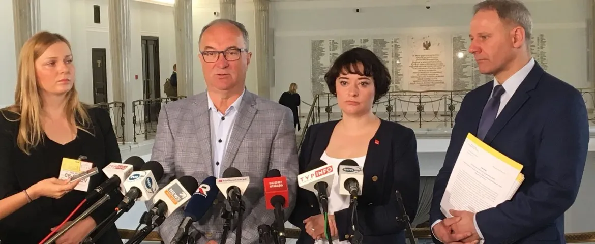 Cztery osoby stoją za zestawem mikrofonów podczas konferencji prasowej w wnętrzu budynku parlamentu, omawiającej koszty finansowania kościołów.