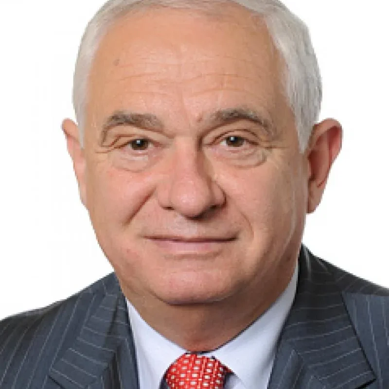 Portret starszego mężczyzny w ciemnym garniturze z czerwonym krawatem na białym tle.