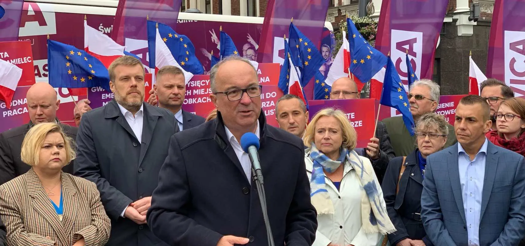 Mężczyzna z okularami przemawia do mikrofonu podczas wiecu lewicowego, w tle tłum ludzi, flagi UE i Polski oraz kolorowe banery.