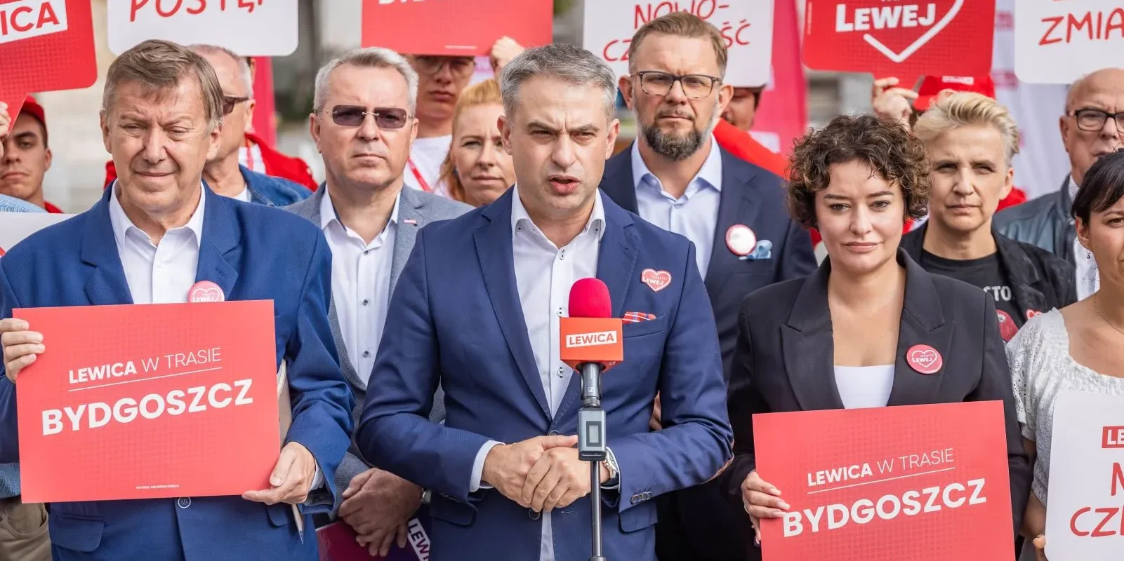 Grupa polityków Lewicy podczas konferencji prasowej, trzymająca czerwone plakaty z napisem „LEWICA W TRASIE BYDGOSZCZ” oraz mikrofon z logiem Lewicy.