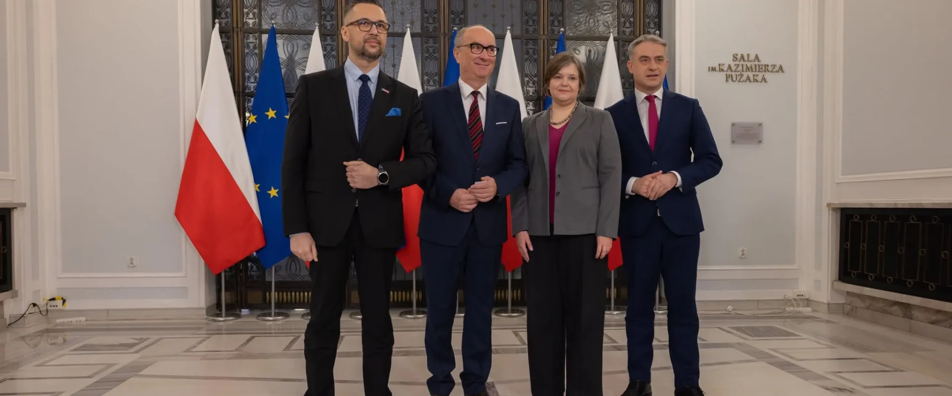 Grupa czterech osób w garniturach stoi razem w eleganckiej sali z flagami Polski i Unii Europejskiej w tle.
