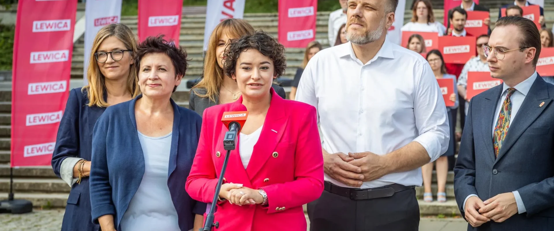 Grupa polityków Lewicy na konferencji prasowej na schodach; kobieta w różowym garniturze przemawia do mikrofonu z logo Lewica, w tle czerwone banery z napisem Lewica.