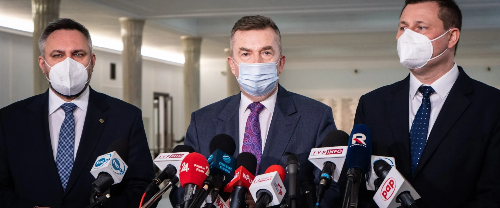 Trzej mężczyźni w maskach ochronnych stoją przed mikrofonami podczas konferencji prasowej w jasnym wnętrzu.