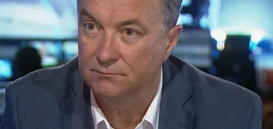 Zbliżenie na mężczyznę w średnim wieku w granatowej marynarce i białej koszuli, siedzącego w studiu telewizyjnym z poważnym wyrazem twarzy.