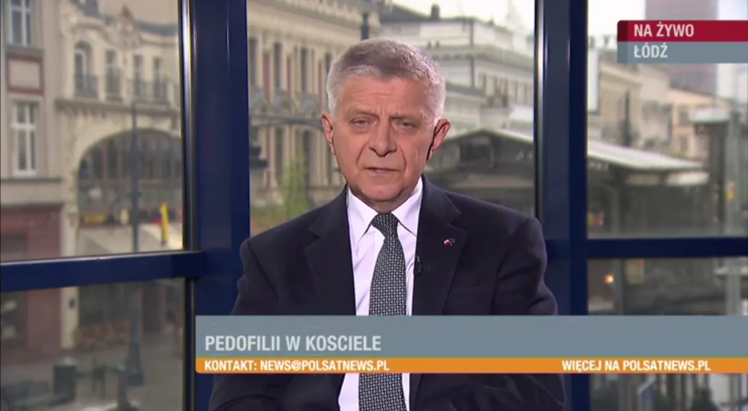 Mężczyzna w garniturze prowadzi program informacyjny w studiu telewizyjnym; na dole ekranu widnieje pasek z napisem „PEDOFILII W KOŚCIELE” i adresem kontaktowym, a w tle widać miejskie budynki.