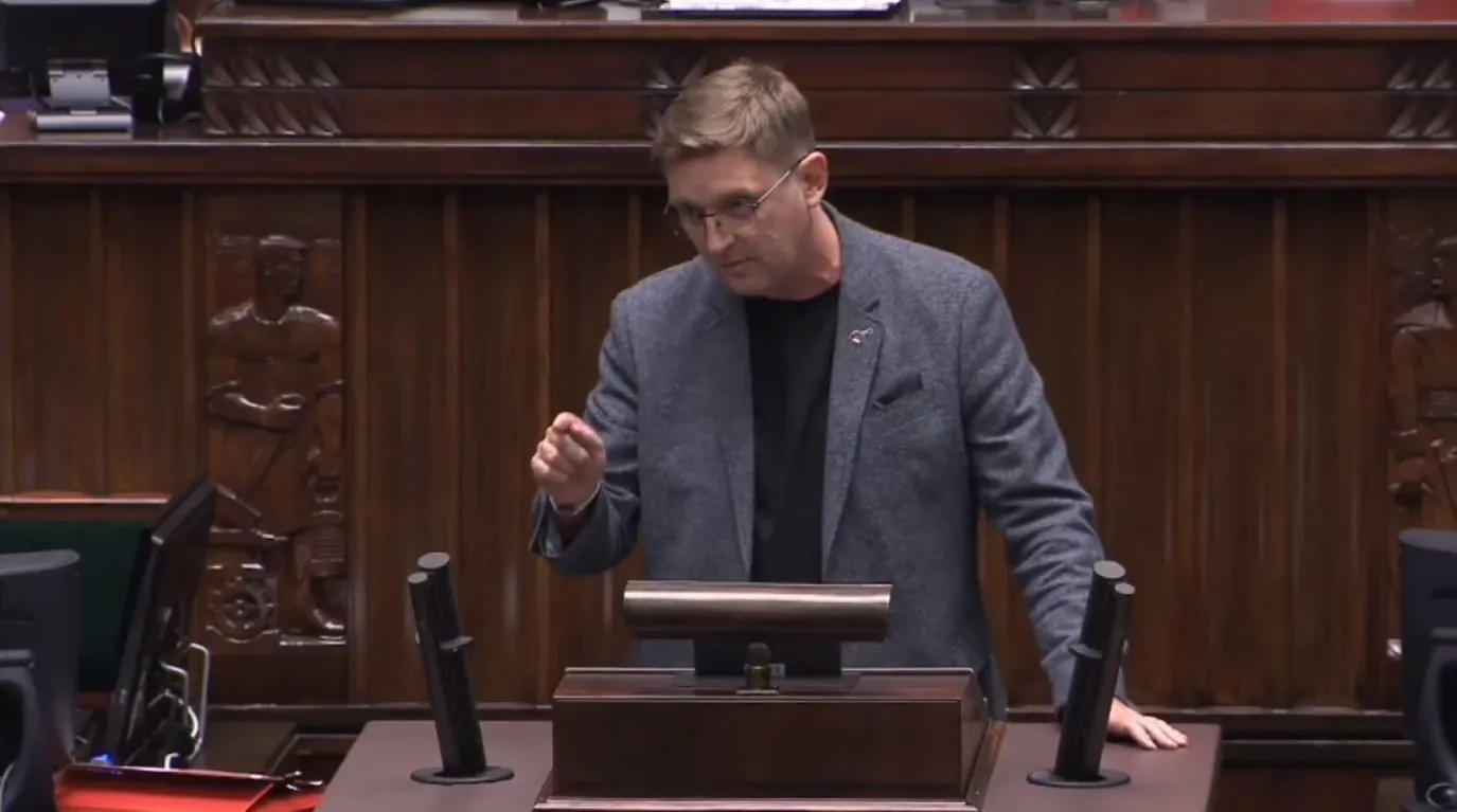 Mężczyzna w szarym żakiecie i czarnej koszuli stoi przy mównicy w sali plenarnej, gestykulując prawą dłonią.