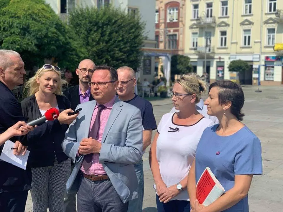 Grupa osób na placu prowadzi konferencję prasową; mężczyzna w jasnym garniturze i różowej koszuli wygłasza oświadczenie do mikrofonów, a obok stoją dwie kobiety z notatkami.