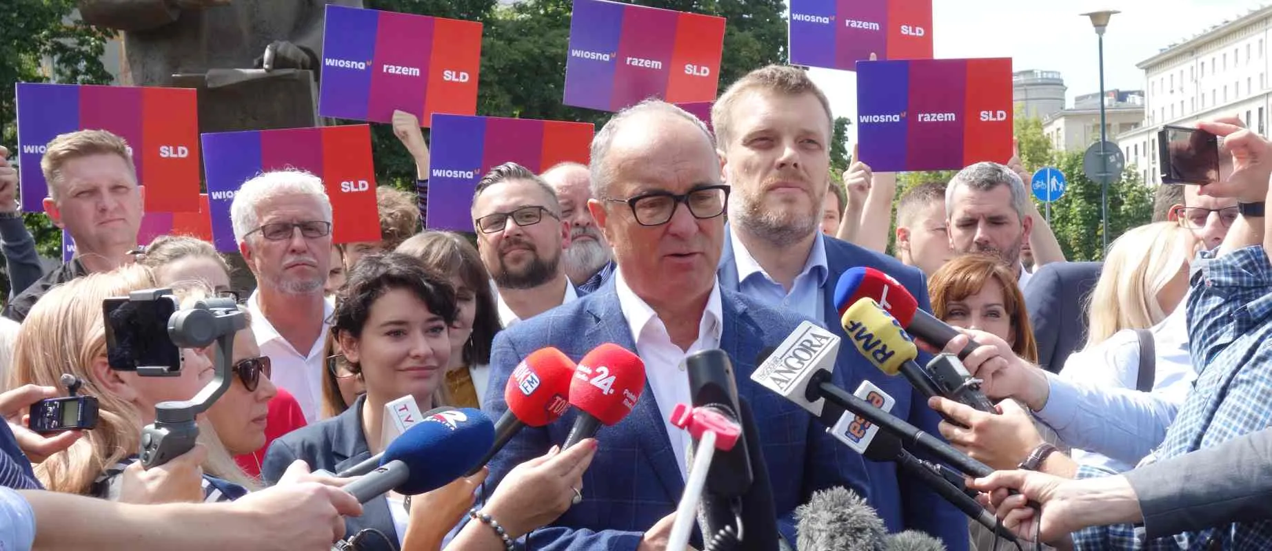 Mężczyzna w granatowym garniturze przemawia do mediów podczas konferencji prasowej Lewicy, otoczony dziennikarzami z mikrofonami, a w tle widoczne kolorowe plakaty z hasłami Wiosna, Razem i SLD.