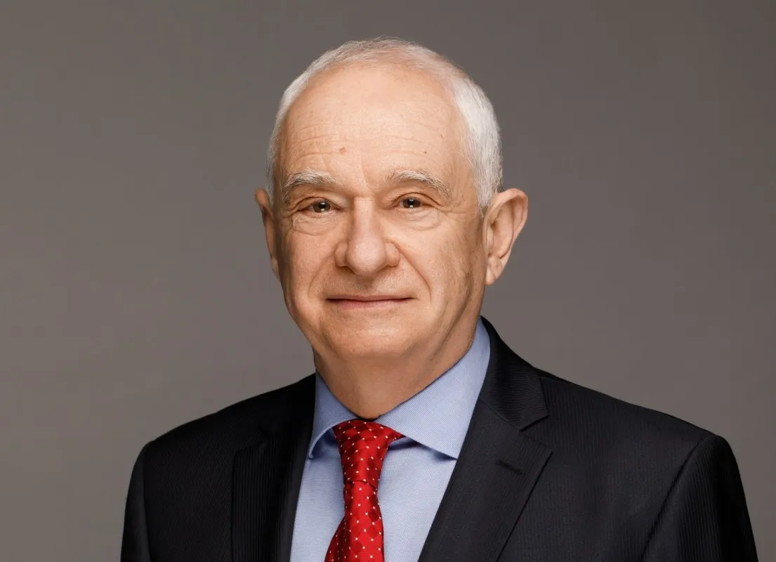 Portret mężczyzny w czarnym garniturze, niebieskiej koszuli i czerwonym krawacie na neutralnym tle.