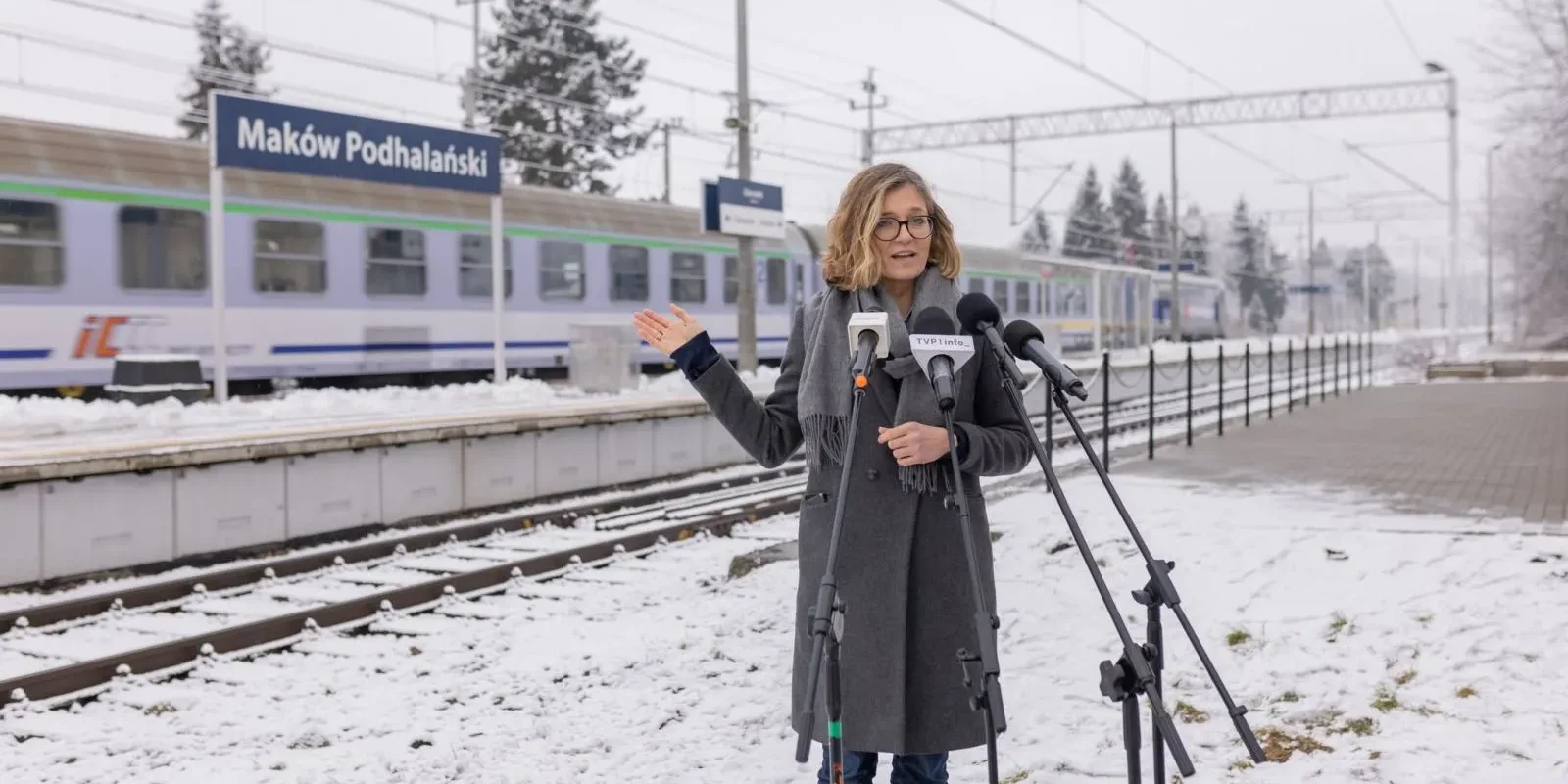 Kobieta w okularach przemawia do mediów z mikrofonami na peronie stacji Maków Podhalański, obok pociągu w zimowej scenerii.