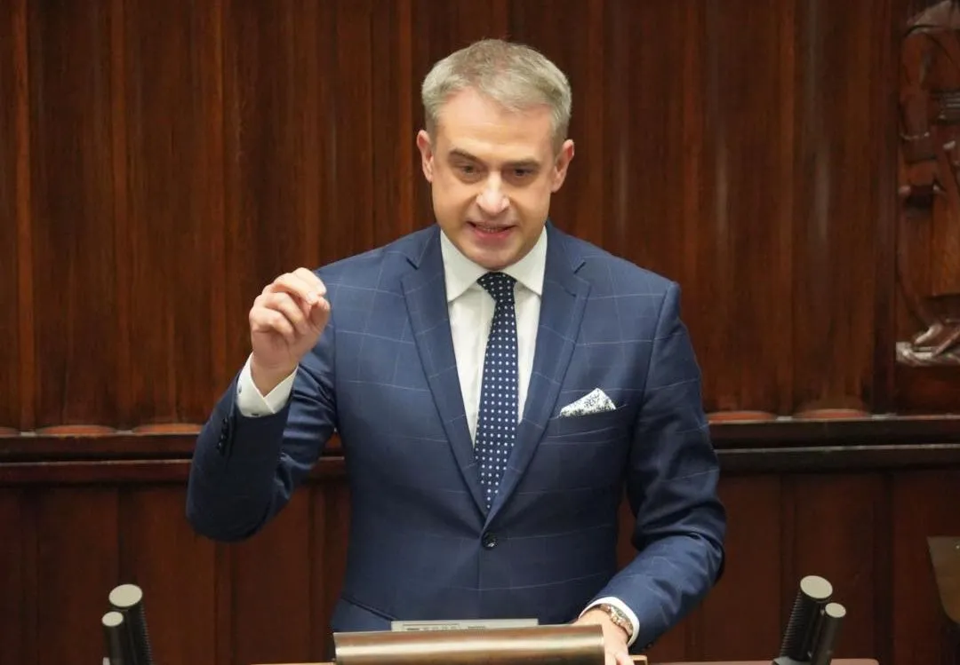 Mężczyzna w granatowym garniturze stoi przy mównicy w sali plenarnej z drewnianymi panelami, gestyklując dłonią.