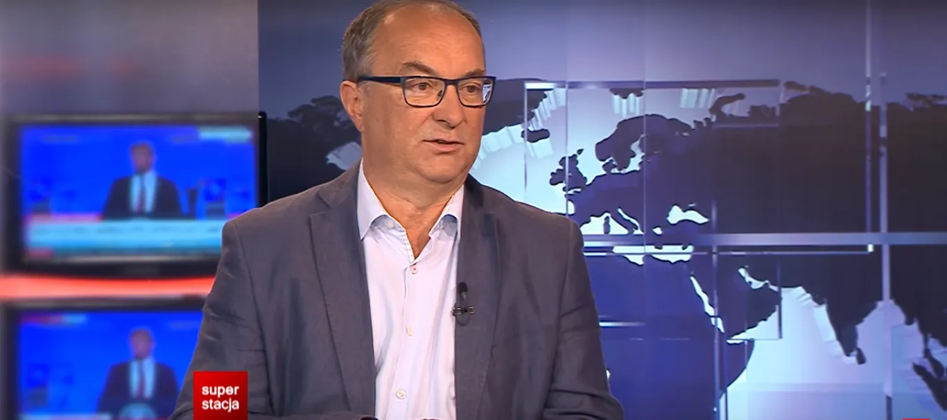 Mężczyzna w garniturze i jasnej koszuli z mikrofonem na klipsie siedzi w studiu telewizyjnym, a w tle widać mapę świata.