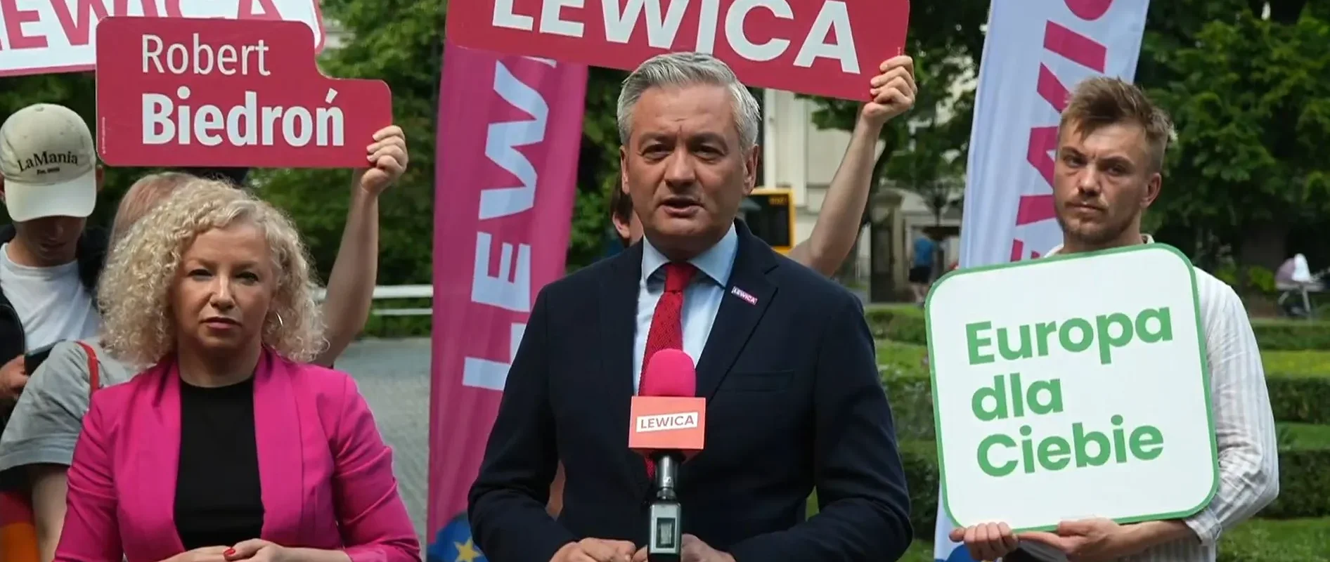 Osoby na konferencji prasowej trzymają banery z napisami Lewica i Europa dla Ciebie, podczas gdy mężczyzna w garniturze przemawia do mikrofonu.