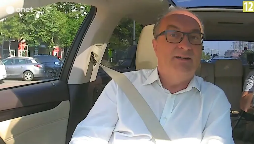 Mężczyzna w białej koszuli i okularach siedzi w samochodzie z zapiętym pasem bezpieczeństwa.