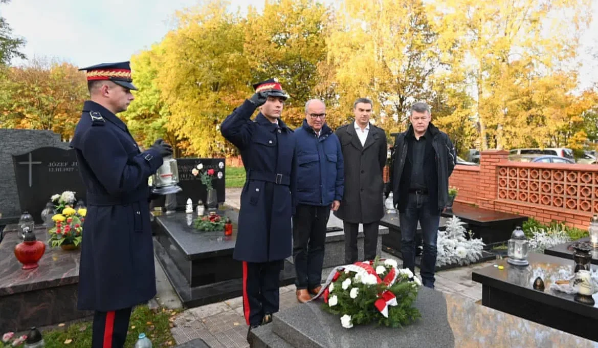 Na cmentarzu w jesiennej scenerii grupa ludzi wraz z dwoma żołnierzami w mundurach salutuje przy grobie ozdobionym wieńcem kwiatów — Pamiętamy.