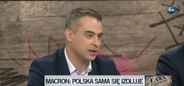 Panelista w garniturze przemawia w studiu telewizyjnym; na dole ekranu widnieje napis "MACRON: POLSKA SAMA SIE IZOLUJE".