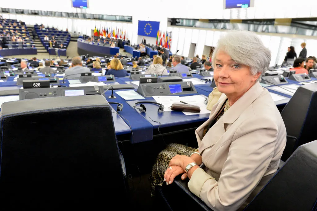 Kobieta w beżowym garniturze siedzi przy stanowisku w sali plenarnej Parlamentu Europejskiego, z flagą UE w tle.