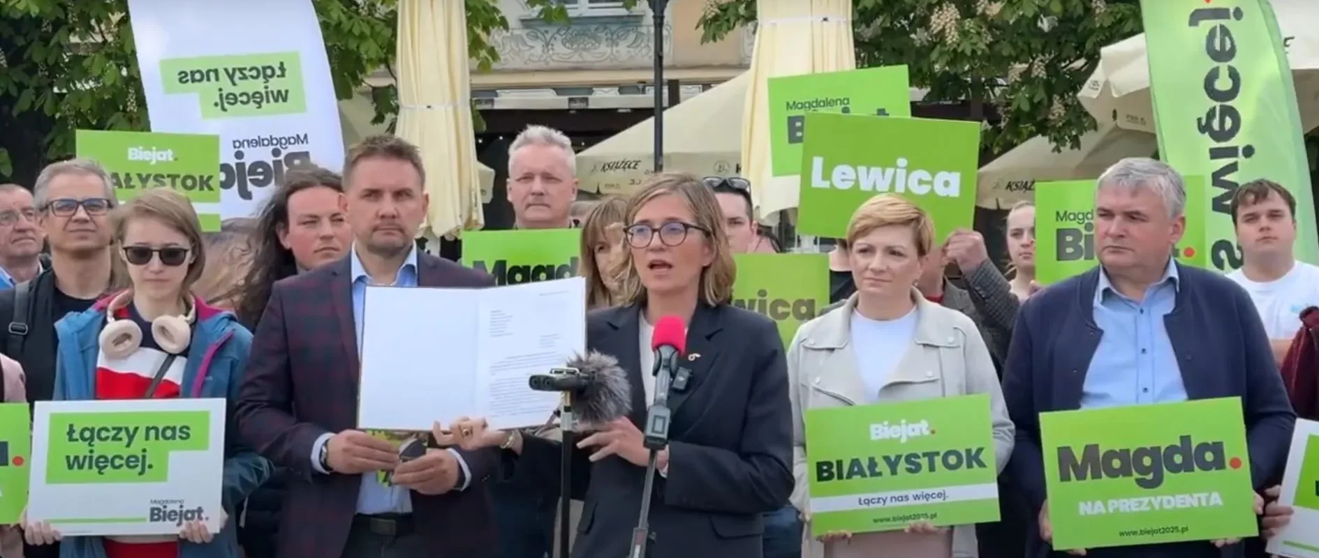 Kobieta w garniturze przemawia do mikrofonu podczas konferencji prasowej, otoczona grupą ludzi z zielonymi plakatami i dokumentami, w kontekście składania doniesienia do prokuratury na Grzegorza Brauna.