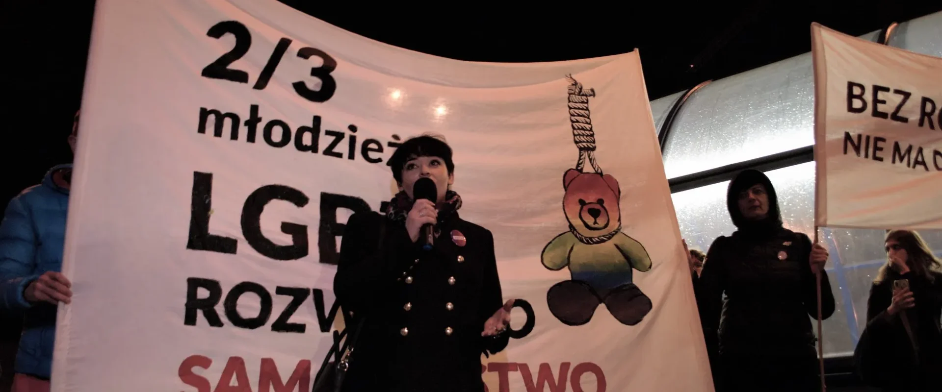 Kobieta przemawia do mikrofonu podczas wieczornego protestu; w tle widać duże banery z hasłami i rysunkiem misia.