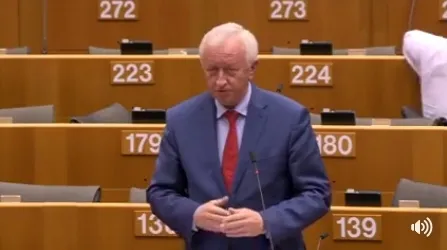Mężczyzna w granatowym garniturze i czerwonym krawacie stoi i przemawia w sali obrad z numerowanymi miejscami, podczas debaty o ochronie osób dorosłych szczególnej troski.