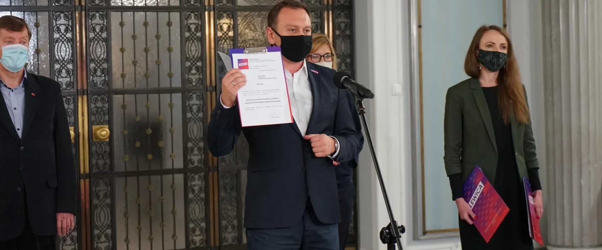 Grupa polityków Lewicy podczas konferencji prasowej przed wejściem do budynku z ozdobnymi drzwiami; centralnie stojący mężczyzna w maseczce trzyma dokumenty przy mikrofonie, a po jego obu stronach stoją współpracownicy także w maseczkach.