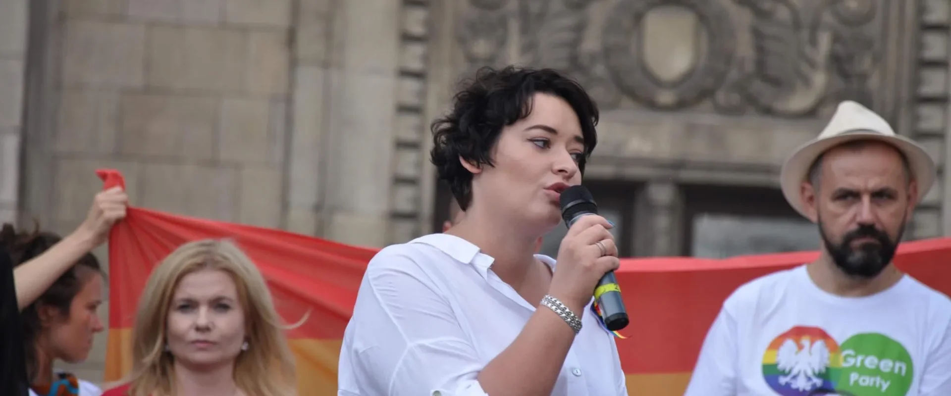 Kobieta z krótkimi czarnymi włosami przemawia do mikrofonu podczas demonstracji, na tle tęczowej flagi i stojących obok uczestników.