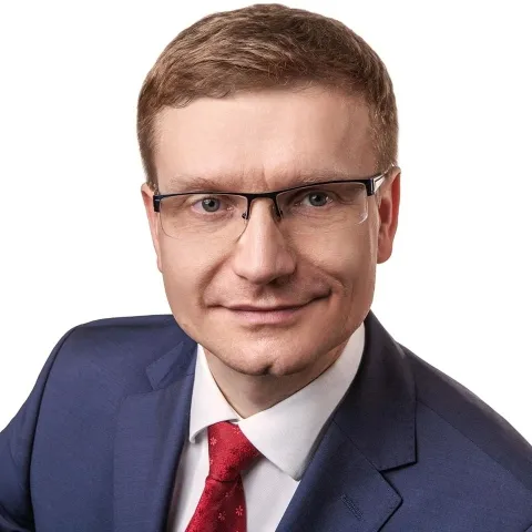 Portret mężczyzny w granatowym garniturze, białej koszuli i czerwonym krawacie, z okularami, na białym tle, uśmiechnięty.
