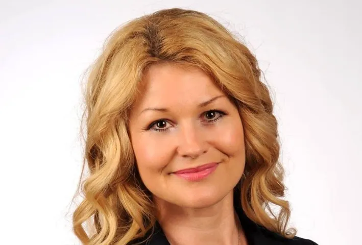 Portret uśmiechniętej kobiety o blond włosach na białym tle, ubranej w czarną bluzkę.