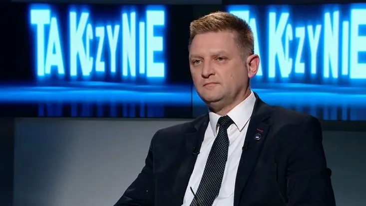 Mężczyzna w garniturze siedzi w studiu telewizyjnym, na tle widoczne są niebieskie napisy.