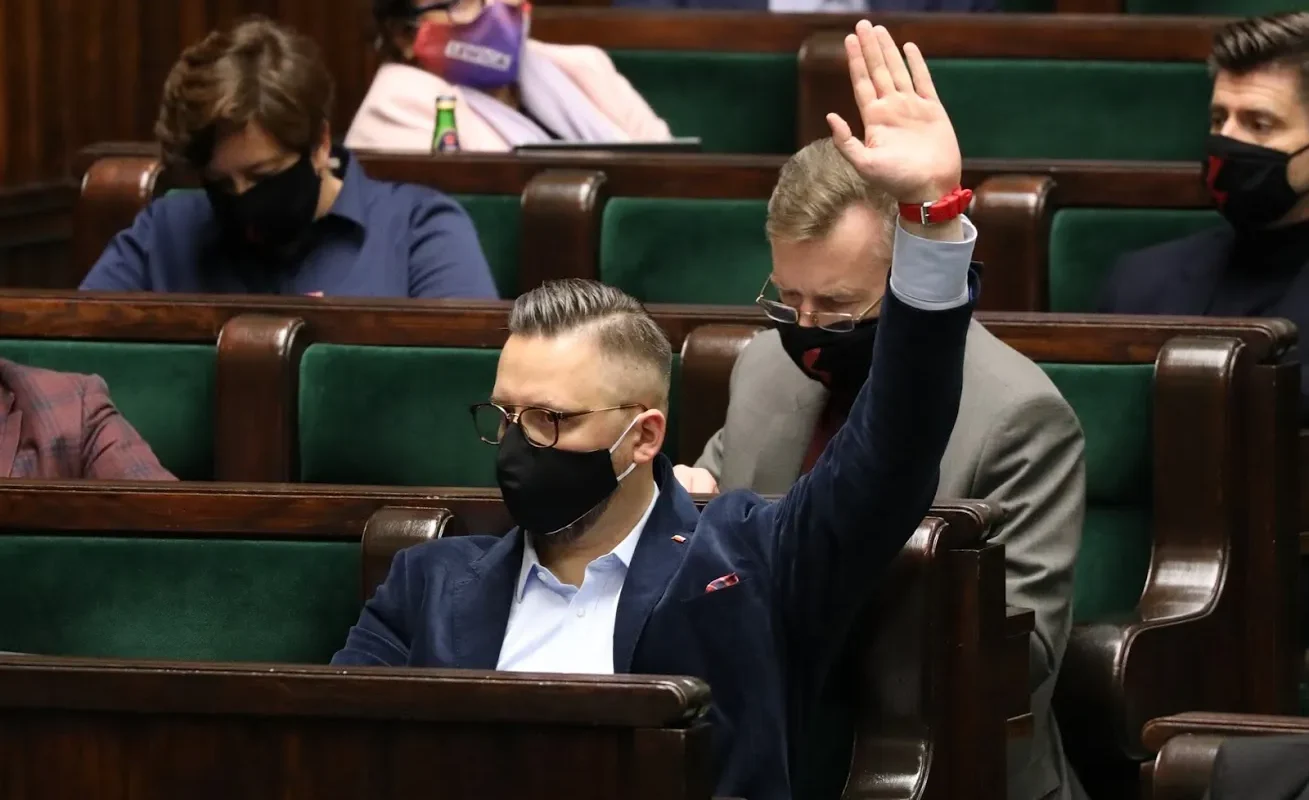 Członek parlamentu w maseczce i garniturze unosi rękę podczas posiedzenia w sali plenarnej, w otoczeniu innych posłów.