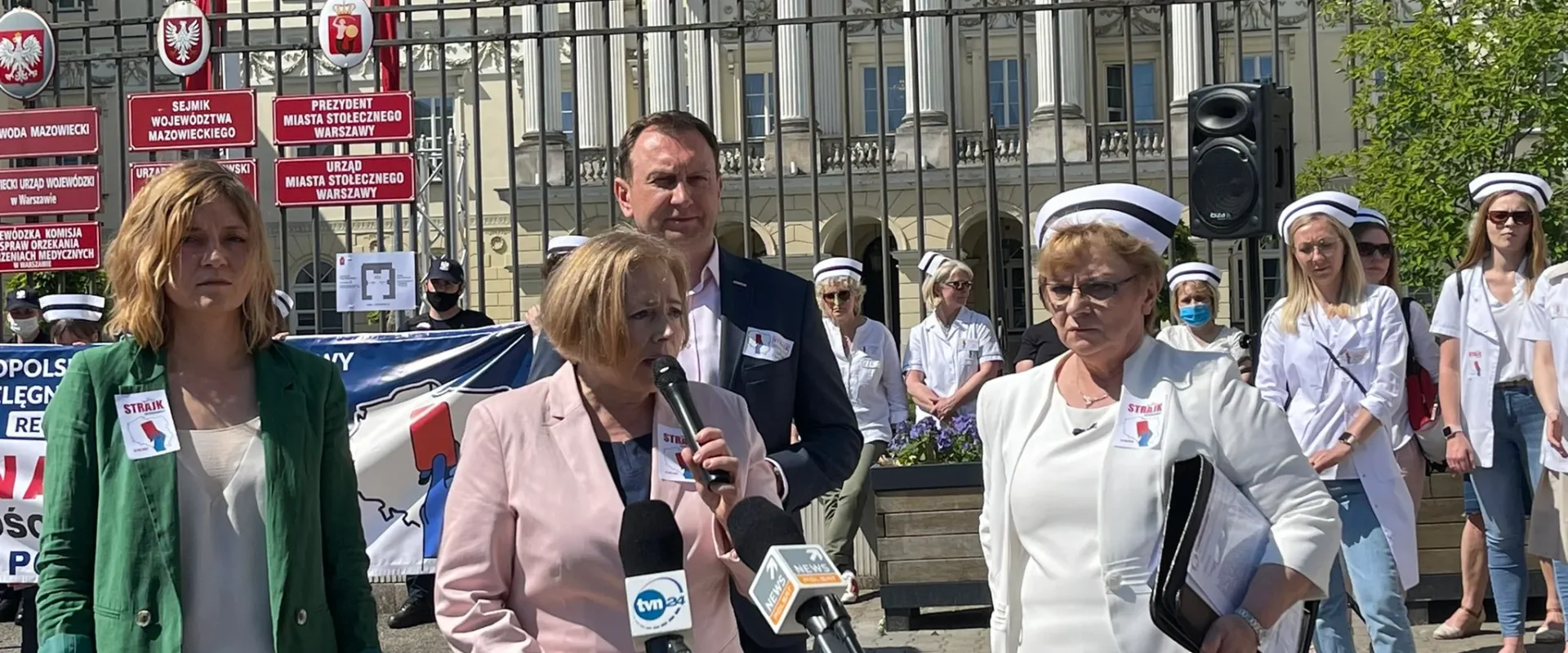 Grupa pielęgniarek i położnych w białych fartuchach i czapkach pielęgniarskich stoi przed kolumnowym budynkiem z metalową balustradą i wygłasza przemówienie podczas ostrzegawczego strajku w Warszawie.