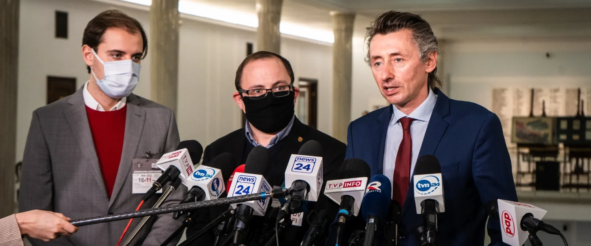 Na zdjęciu trzech mężczyzn stoi przed mikrofonami podczas konferencji prasowej w budynku, prawdopodobnie rządowym.