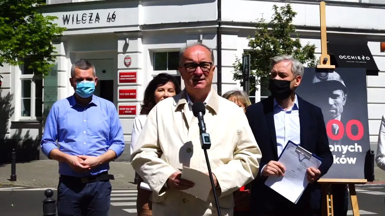 Grupa ludzi stojących na zewnątrz budynku, centralny mężczyzna w jasnym płaszczu przemawia do mikrofonu i trzyma kartkę, obok niego inni z dokumentami, w tle plakat i wejście do budynku.