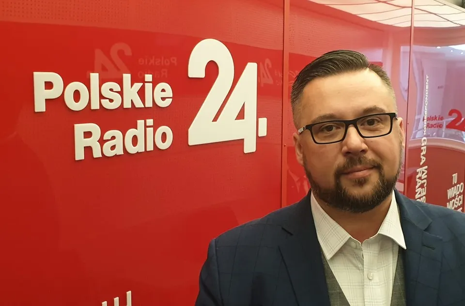 Mężczyzna z brodą i okularami w granatowym garniturze stoi na tle czerwonej ściany z napisem „Polskie Radio 24”.
