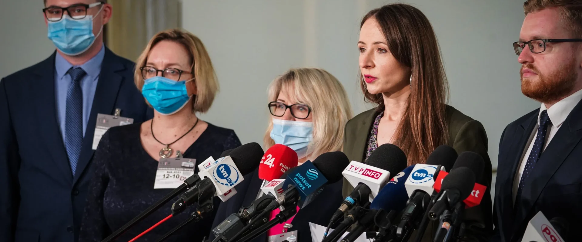 Na zdjęciu przedstawiono grupę czterech osób w maseczkach ochronnych, stojących przy mikrofonach podczas konferencji prasowej.