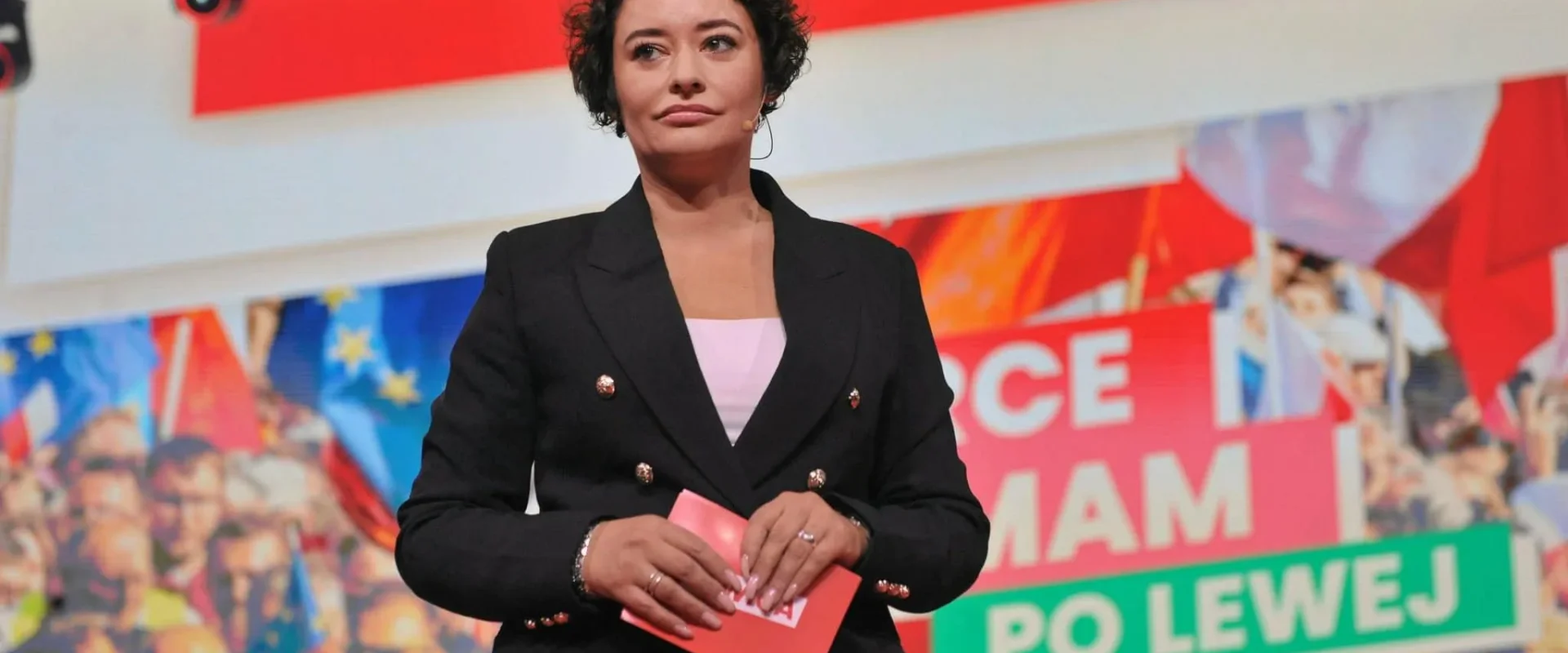 Kobieta ubrana w czarny żakiet stoi na scenie, trzyma różowy notes, a w tle widać kolorowe banery i zgromadzonych uczestników; kontekst dotyczy wypowiedzi o obniżce składki zdrowotnej.