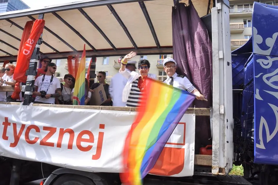 Uczestnicy Parady Równości stoją na platformie ciężarówki, machają tęczową flagą i pozują do zdjęć podczas parady.
