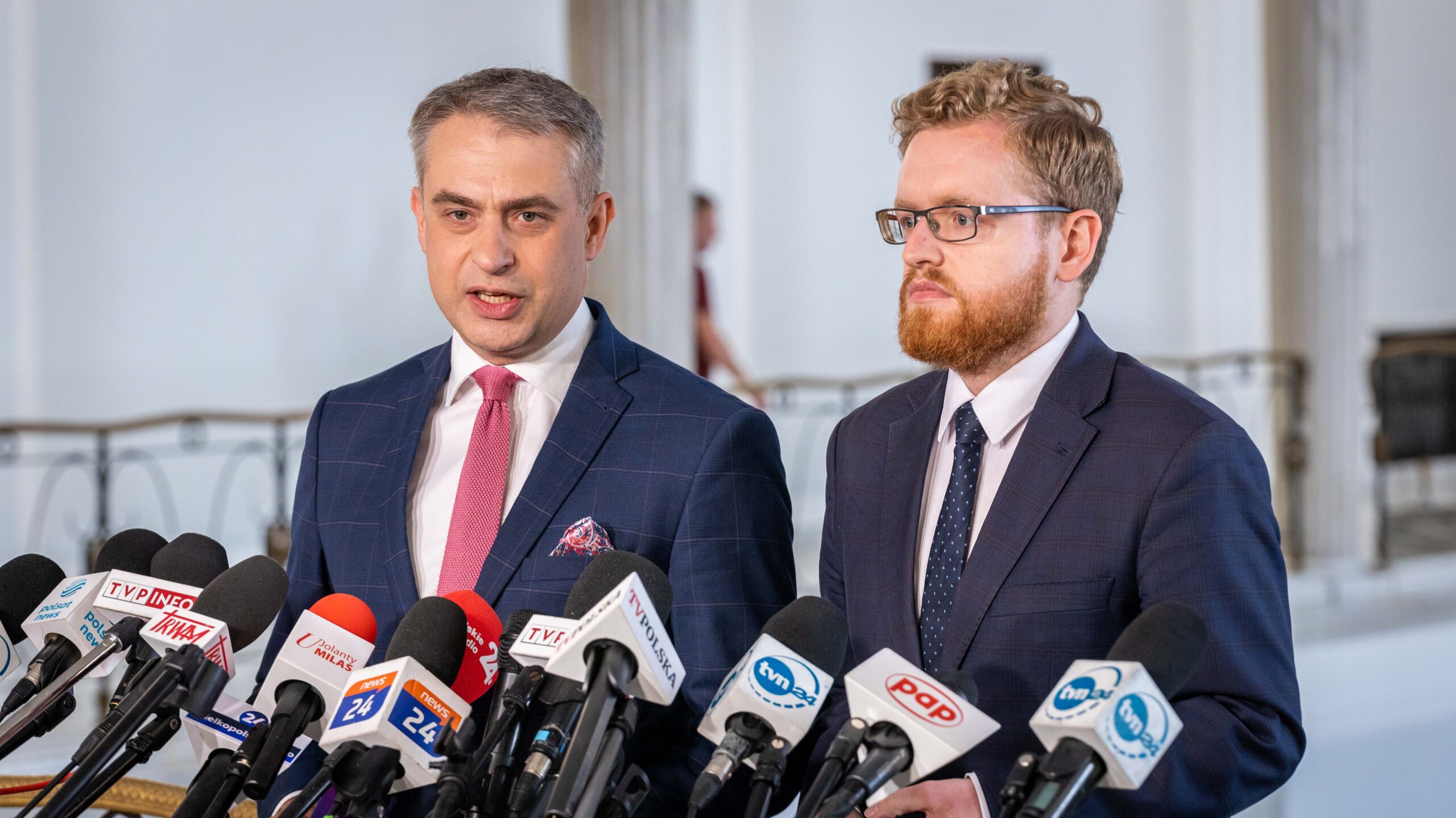 Dwóch mężczyzn w garniturach udziela konferencji prasowej, stojąc przed licznymi mikrofonami z logotypami stacji telewizyjnych.