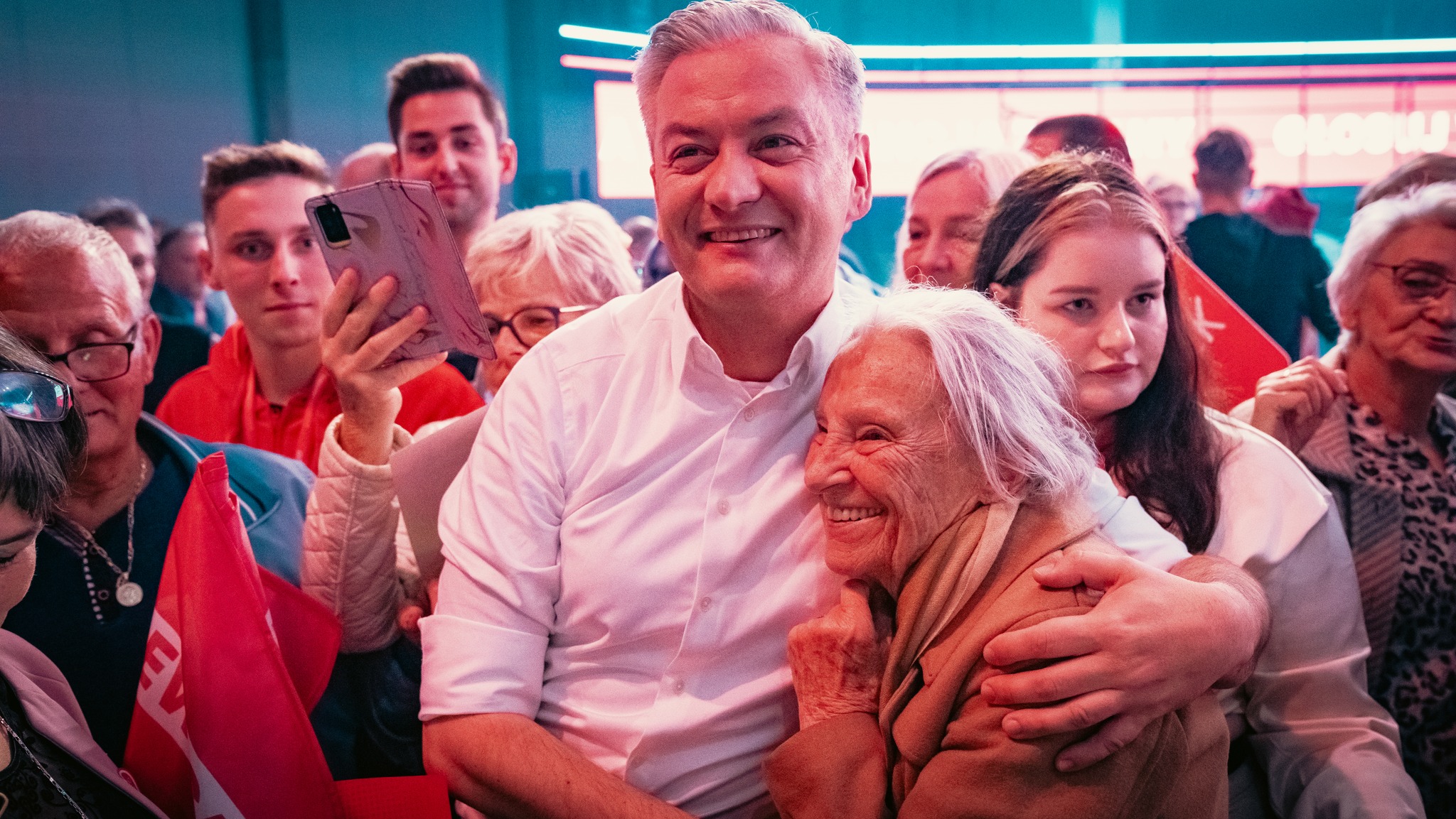 Mężczyzna w białej koszuli przytula starszą kobietę w tłumie podczas konwencji w Będzinie, wokół niego ludzie trzymają czerwone flagi.