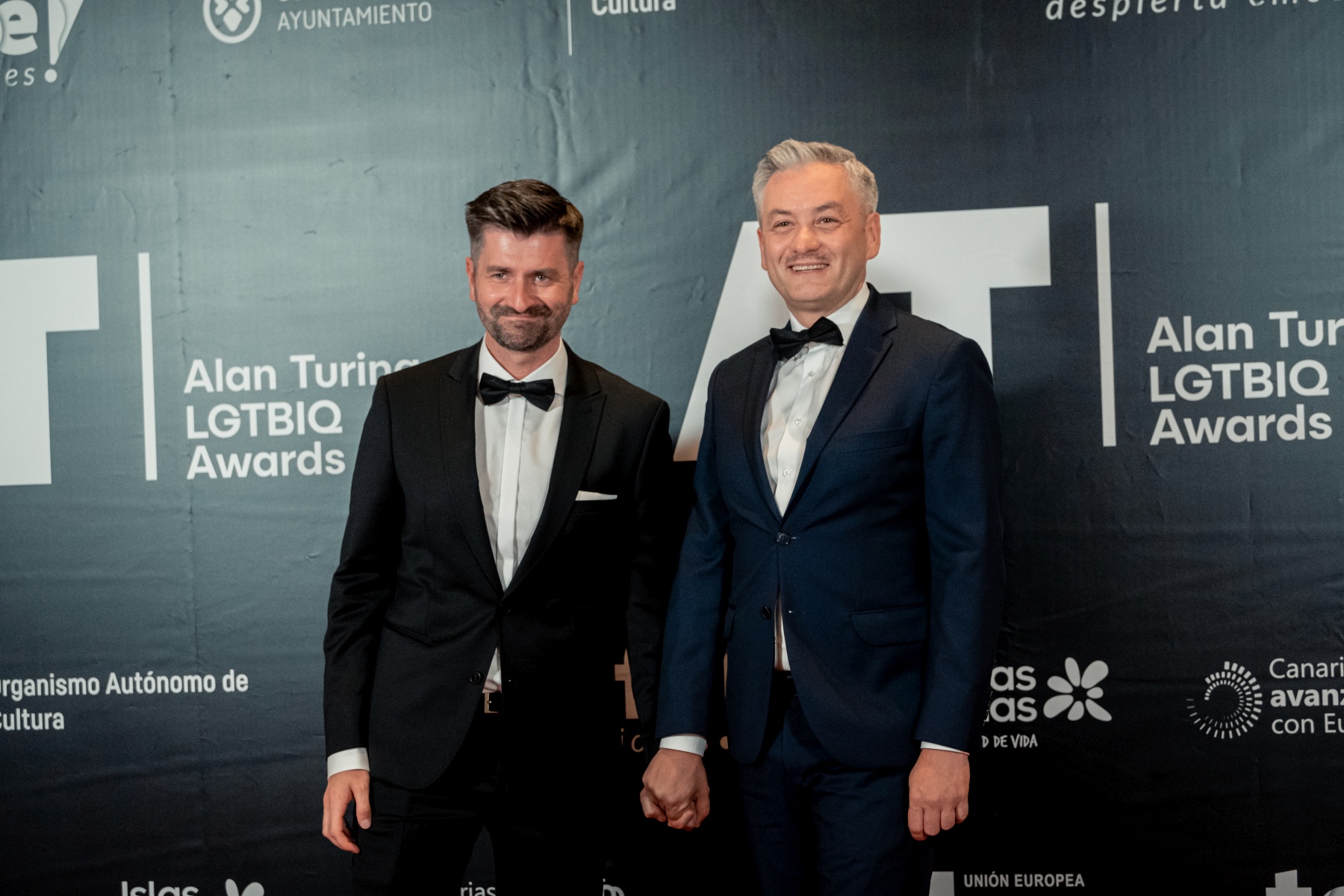 Dwóch mężczyzn ubranych w smokingi stoi na tle ścianki z logo Alan Turing LGBTQ+ Awards, trzymają się za ręce i uśmiechają.