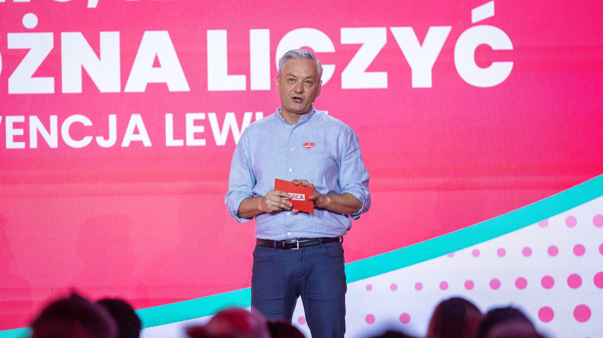 Mężczyzna w jasnoniebieskiej koszuli stoi na scenie, trzyma czerwony notes, a w tle widać duży różowy ekran z białymi napisami.